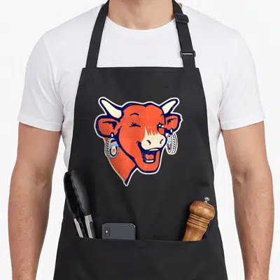 Tablier de cuisine noir avec tête de vache orange souriante portant des boucles d'oreilles en pots de fromage stylisées.