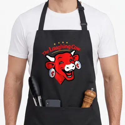 Tablier de cuisine noir avec tête de vache rouge souriante et texte The Laughing Cow en rouge.