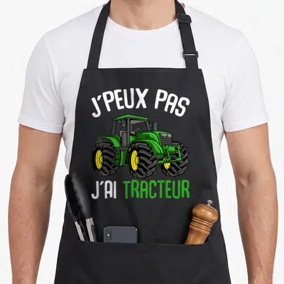 Tablier de cuisine noir avec illustration d'un tracteur vert et du texte J'peux pas j'ai tracteur en blanc et vert.