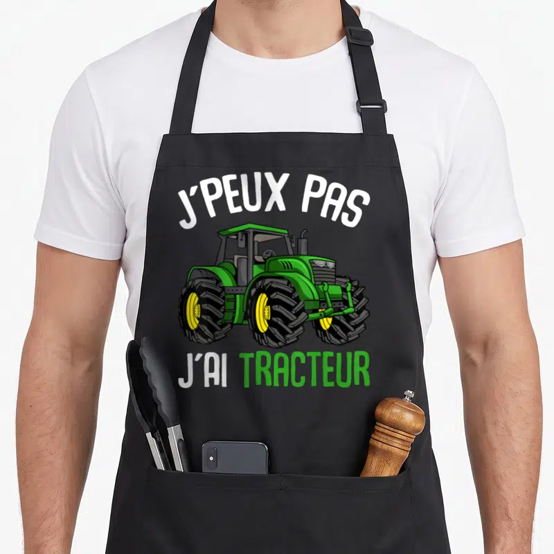 Tablier de cuisine noir avec illustration d'un tracteur vert et du texte J'peux pas j'ai tracteur en blanc et vert.