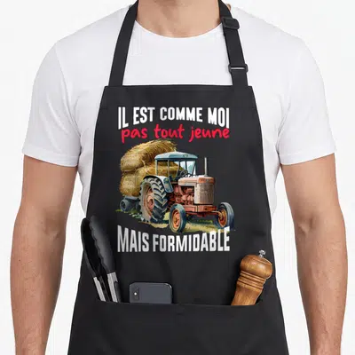 Tablier de cuisine noir avec illustration d'un vieux tracteur orange tirant une remorque chargée de foin, entouré du texte Il est comme moi pas tout jeune mais formidable.