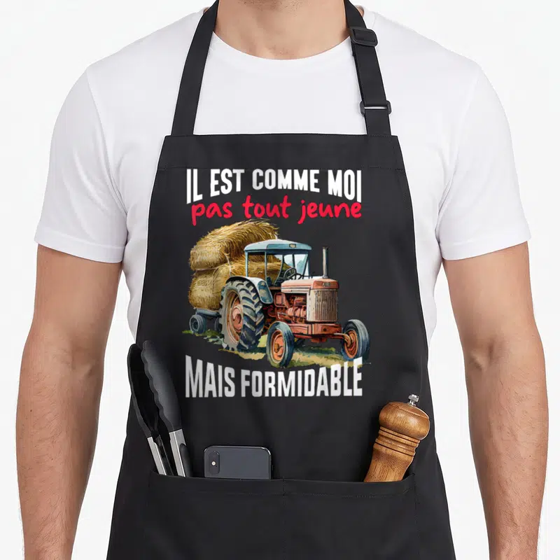 Tablier de cuisine noir avec illustration d'un vieux tracteur orange tirant une remorque chargée de foin, entouré du texte Il est comme moi pas tout jeune mais formidable.