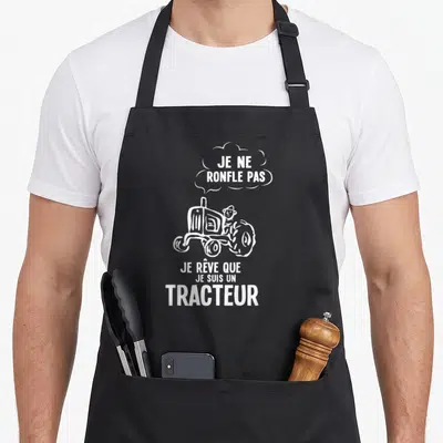 Tablier de cuisine noir avec illustration blanche d'un tracteur et texte humoristique sur fond uni.