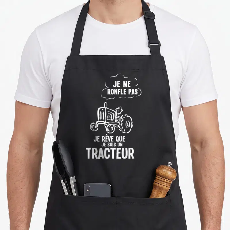 Tablier de cuisine noir avec illustration blanche d'un tracteur et texte humoristique sur fond uni.