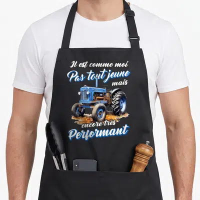 Tablier de cuisine noir avec illustration d'un tracteur bleu et texte blanc Il est comme moi pas tout jeune mais encore très performant.