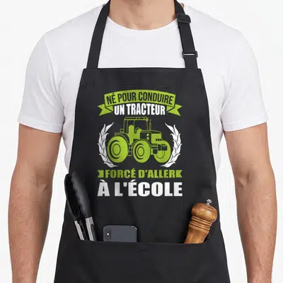 Tablier de cuisine noir avec illustration verte d'un tracteur encadré de lauriers blancs et texte humoristique sur le métier agricole.