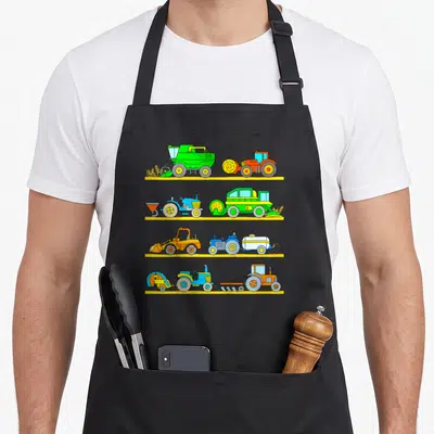 Tablier de cuisine noir avec quatre rangées de tracteurs, moissonneuses et batteuses colorés sur fond jaune.