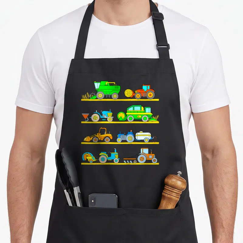 Tablier de cuisine noir avec quatre rangées de tracteurs, moissonneuses et batteuses colorés sur fond jaune.