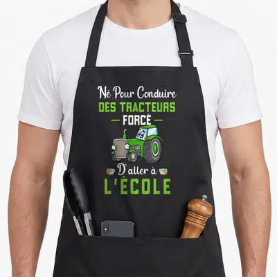 Tablier de cuisine noir avec illustration d'un tracteur vert et le texte Né Pour Conduire Des Tracteurs Forcé D'aller à L'école