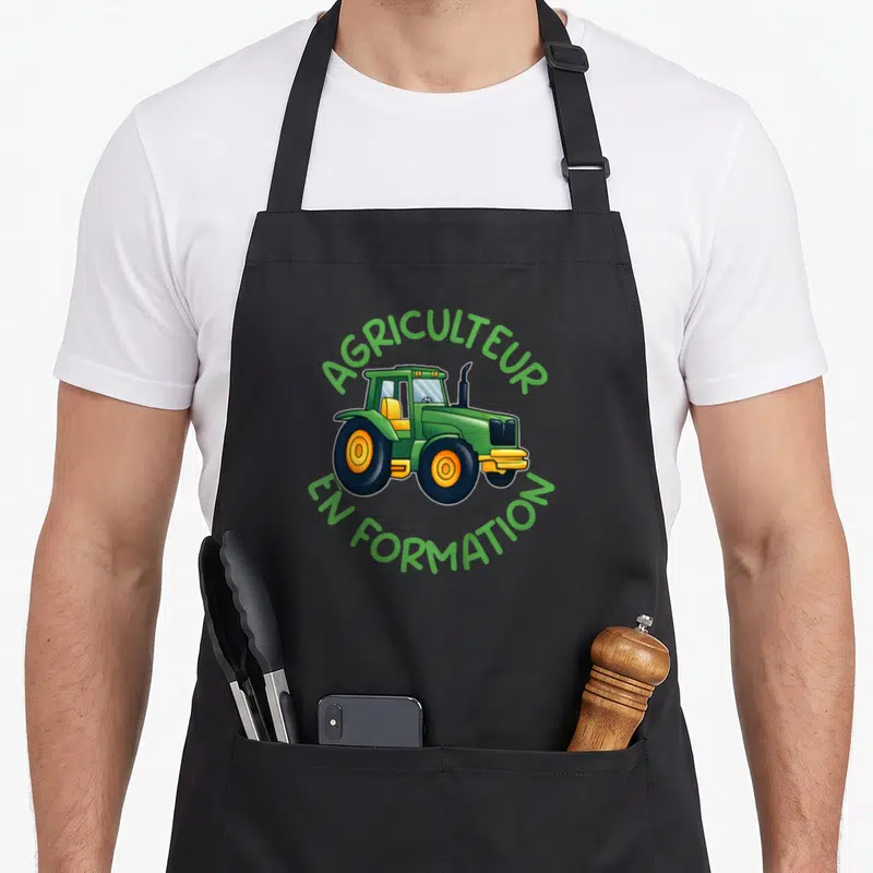 Tablier de cuisine noir avec illustration de tracteur vert et texte agricultural en formation en cercle.