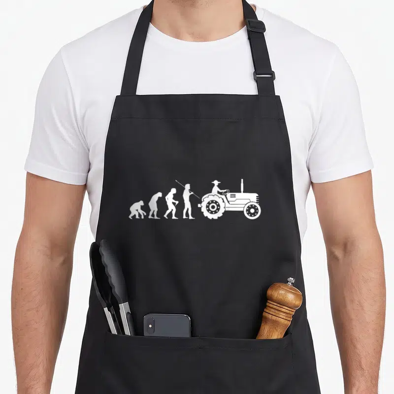 Tablier de cuisine noir avec silhouette blanche montrant l'évolution humaine vers un tracteur agricole.
