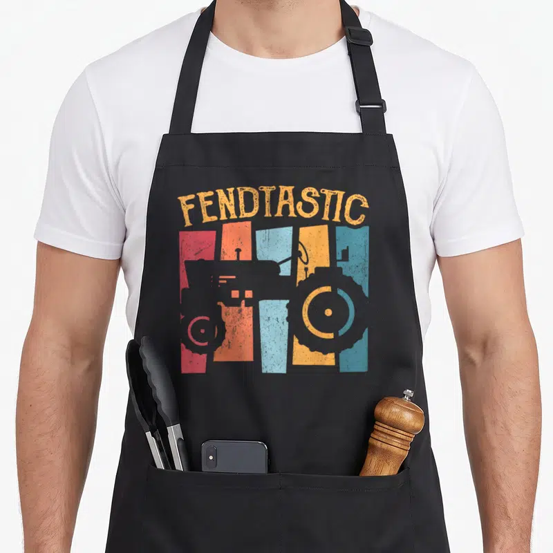 Tablier de cuisine noir avec illustration de tracteur stylisé sur bandes colorées et texte Fendtastic en orange.