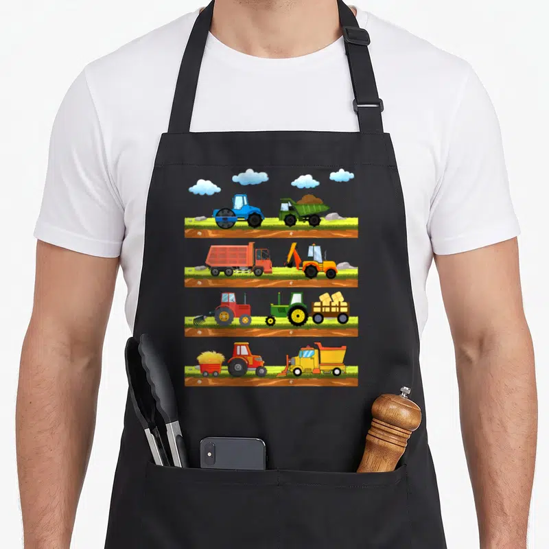 Tablier de cuisine noir avec quatre bandes illustrant des tracteurs et engins de chantier colorés sur un terrain terreux et verdoyant sous des nuages.