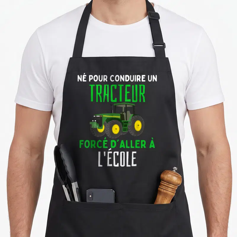 Tablier de cuisine noir avec illustration d'un tracteur vert et jaune et texte humoristique sur fond uni.