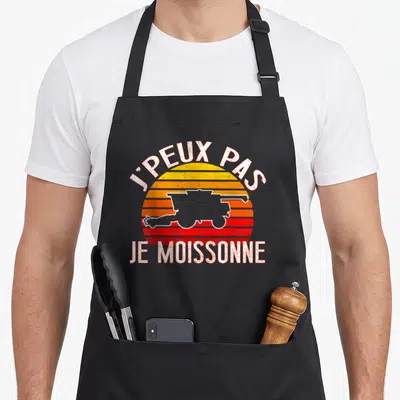 Tablier de cuisine noir avec motif rétro J'peux pas je moissonne et silhouette de tracteur sur fond de soleil orange et rouge.