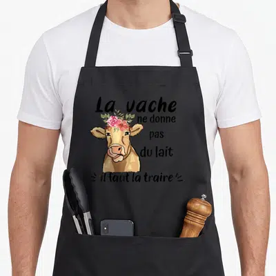 Tablier de cuisine noir avec illustration d'une vache aux fleurs et le texte humoristique sur la production de lait.