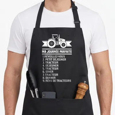 Tablier de cuisine noir avec illustration de tracteur et texte blanc listant une journée idéale centrée sur la machine agricole.