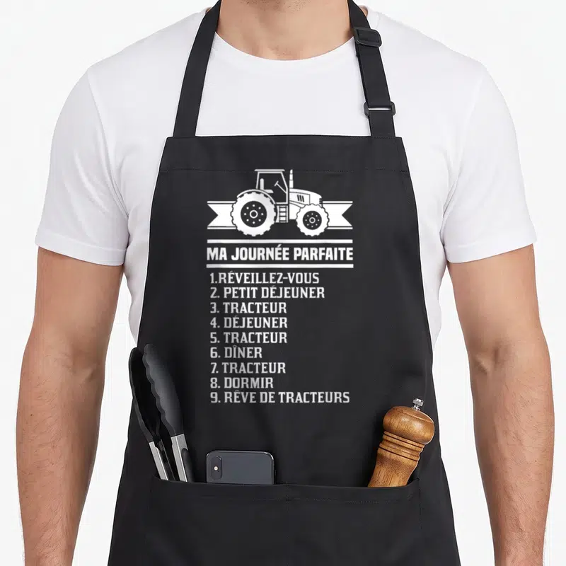 Tablier de cuisine noir avec illustration de tracteur et texte blanc listant une journée idéale centrée sur la machine agricole.