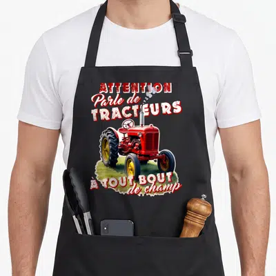 Tablier de cuisine noir avec illustration de tracteur rouge ancien et texte humoristique sur fond de verdure.