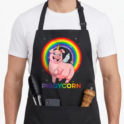 Tablier de cuisine noir avec un cochon licorne rose souriant devant un arc-en-ciel multicolore et l'inscription PIGGYCORN en bas.
