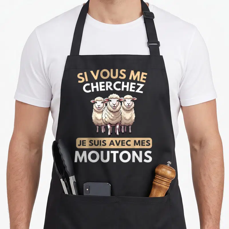 Tablier de cuisine noir avec trois moutons dessinés et le texte Si vous me cherchez je suis avec mes moutons en lettres blanches et dorées.