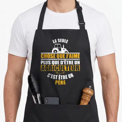 Tablier de cuisine noir avec illustration de tracteur blanc et message jaune sur la paternité agricole.