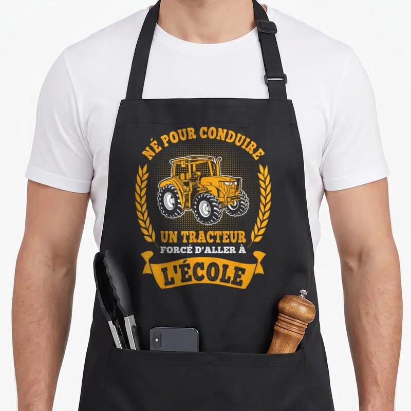 Tablier de cuisine noir avec illustration de tracteur orange encadré par des épis de wheat et le texte né pour conduire un tracteur forcé d'aller à l'école.