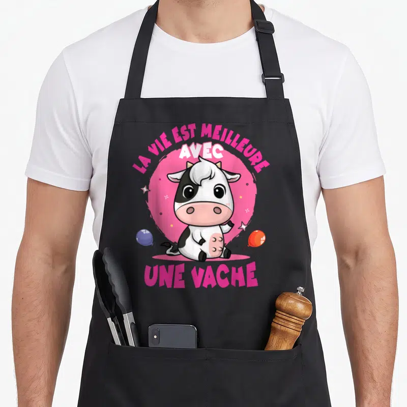 Tablier de cuisine noir avec une illustration de vache mignonne et le texte la vie est meilleure avec une vache en lettres roses.