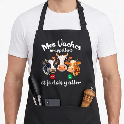 Tablier de cuisine noir avec trois têtes de vaches colorées et texte blanc humoristique sur fond uni.