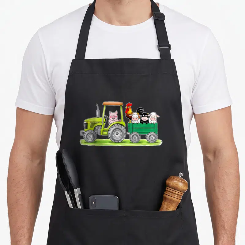 Tablier de cuisine noir avec illustration d'un tracteur vert emmenant un cochon, un coq et des moutons dans une remorque.