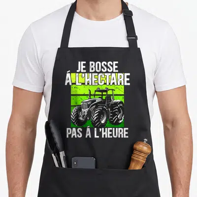 Tablier de cuisine noir avec un tracteur vert et blanc et le texte Je bosse à l'hectare pas à l'heure