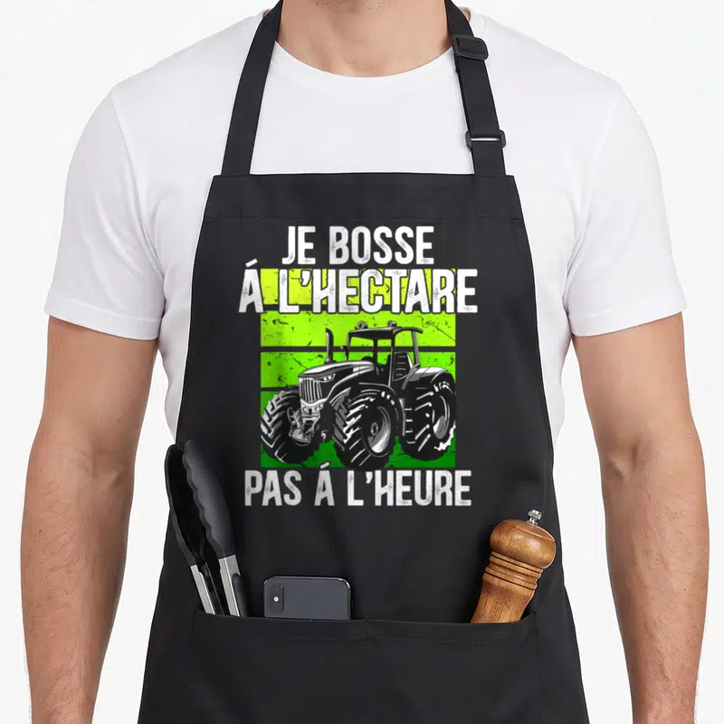 Tablier de cuisine noir avec un tracteur vert et blanc et le texte Je bosse à l'hectare pas à l'heure
