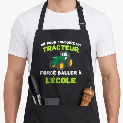 Tablier de cuisine noir avec illustration d'un tracteur vert et texte humoristique sur fond gris uni.