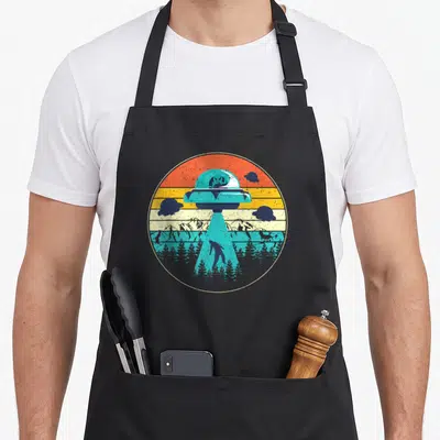 Tablier de cuisine noir avec illustration circulaire d'un ovni turquoise émettant un faisceau lumineux au-dessus d'une forêt.