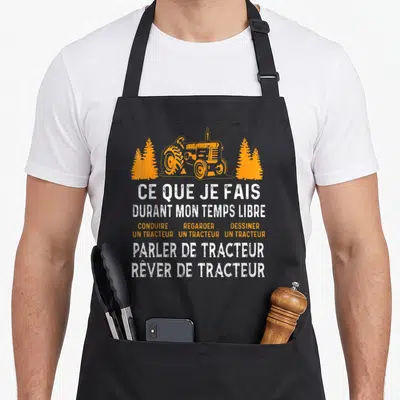 Tablier de cuisine noir avec illustration orange d'un tracteur encadré de pins et texte blanc humoristique sur la passion des engins agricoles.