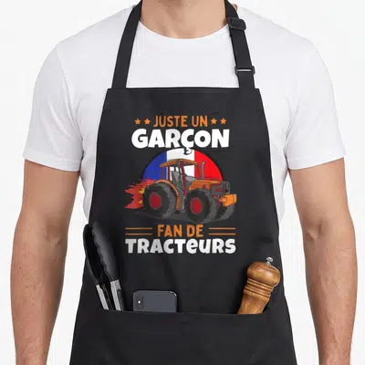 Tablier de cuisine noir avec illustration d'un tracteur orange et du texte Juste Un Garçon Fan De Tracteurs sur fond tricolore.
