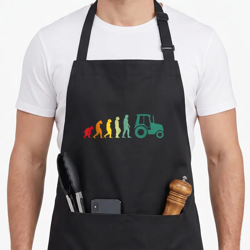 Tablier de cuisine noir avec une illustration colorée montrant l'évolution humaine finissant par un tracteur vert stylisé.