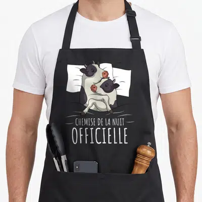 Tablier de cuisine noir avec illustration de deux vaches blotties sous une couverture et le texte « chemise de la nuit » imprimé en blanc.