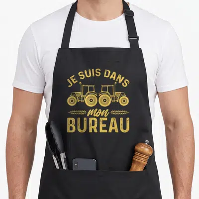 Tablier de cuisine noir avec deux tracteurs jaunes et le texte Je suis dans mon bureau imprimé en centre.