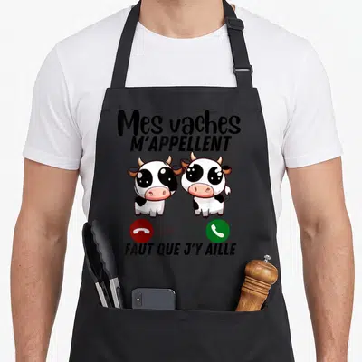 Tablier de cuisine noir avec deux vaches cartoon aux grands yeux et le message Mes vaches m'appellent faut que j'y aille imprimé en noir.