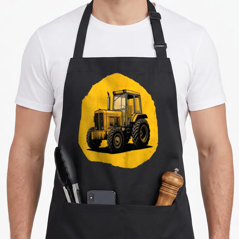 Tablier de cuisine noir avec illustration d'un tracteur jaune sur fond circulaire orange vif.