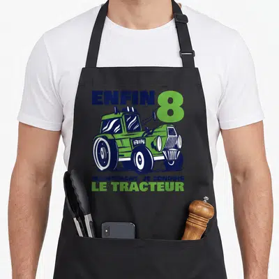 Tablier de cuisine noir avec illustration d'un tracteur vert et texte enfin 8 maintenant je conduis le tracteur en bleu et vert.
