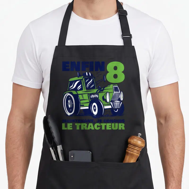 Tablier de cuisine noir avec illustration d'un tracteur vert et texte enfin 8 maintenant je conduis le tracteur en bleu et vert.
