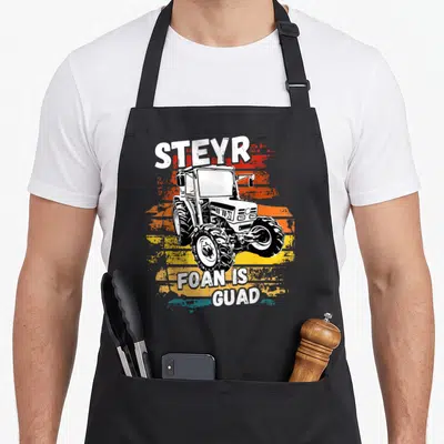 Tablier de cuisine noir avec illustration de tracteur Steyr et texte FOAN IS GUAD sur fond coloré dégradé.