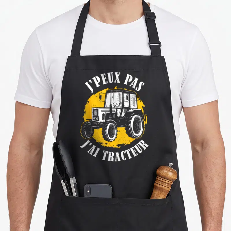 Tablier de cuisine noir avec illustration de tracteur et texte J'peux pas j'ai tracteur en blanc sur fond orange.