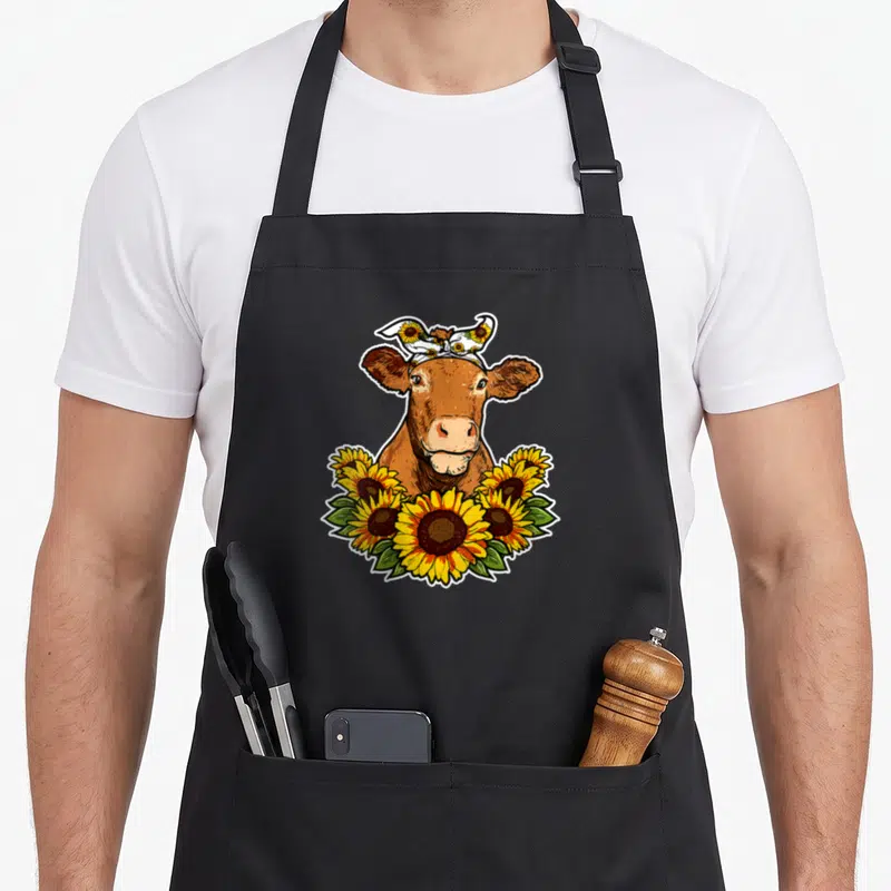 Tablier de cuisine noir avec illustration d'une vache entourée de fleurs de tournesol et d'un foulard floral sur la tête.