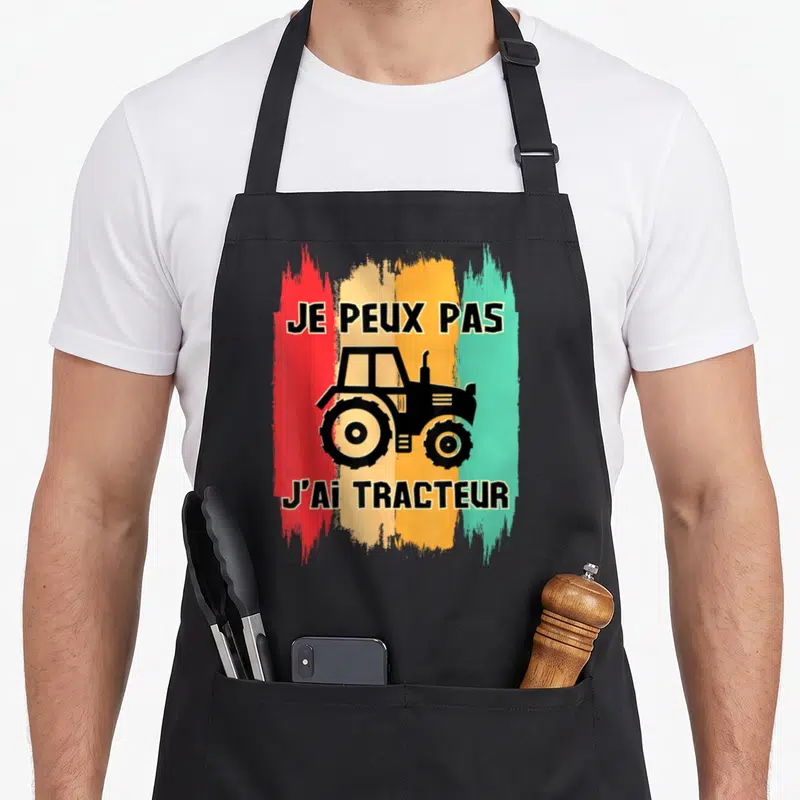 Tablier de cuisine noir avec illustration de tracteur et texte Je peux pas j'ai tracteur sur bandes colorées.