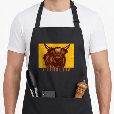 Tablier de cuisine noir avec illustration de vache Highland et texte HIGHLAND COW en orange.