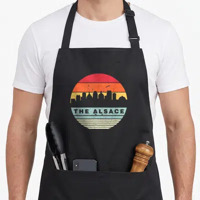 Tablier de cuisine noir avec un motif circulaire coloré représentant une silhouette urbaine et le texte THE ALSACE en lettres capitales grises.
