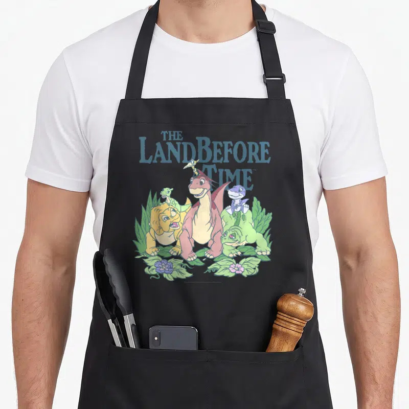 Tablier de cuisine noir avec le logo The Land Before Time et cinq dinosaures pastels entourés de feuilles et de fleurs colorées.